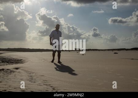 Man in a white shirt walking on the beach with the sunrise behind. Stock Photo