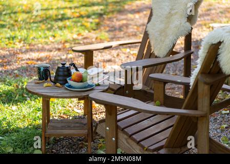 An open book, a kettle with a cup of tea, a plate with apples and pears, on a wooden table with comfortable armchair beside, outdoor in autumn. Stock Photo