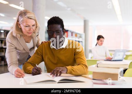 Friendly female tutor helping to diligent african-american man preparing for exam in library Stock Photo