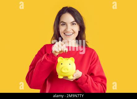 Portrait of happy smiling brunette woman putting coin in yellow piggy bank on yellow background. Stock Photo
