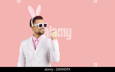 Young handsome man with easter rabbit ears and sunglasses standing over isolated pink background with happy face looking and pointing to the side Stock Photo