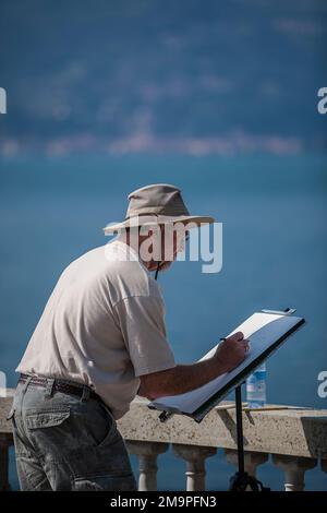 A male American artist, wearing a sun hat and t-shirt, drawing on a terrace overlooking Lake Como in Bellagio, Italy. Stock Photo