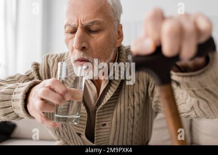 grey haired man with parkinson disease and trembling hands drinking water while sitting with blurred walking cane at home,stock image Stock Photo