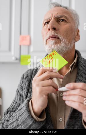 tense man suffering from memory loss while holding sticky notes with phone number and looking away in kitchen,stock image Stock Photo