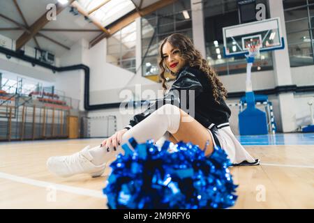 Brunette, curly-haired cheerleader in black and white uniform and jacket sitting on basketball court. Blue shiny pom-pom blurred in the foreground. High quality photo Stock Photo