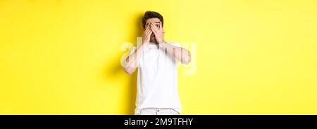 Embarrassed guy shut eyes but peeking through fingers at something awkward, standing over yellow background Stock Photo