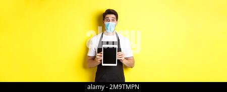 Concept of covid-19, small business and pandemic. Surprised waiter in black apron and medical mask showing tablet screen, standing over yellow Stock Photo