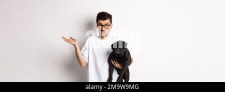 Image of handsome young man holding black pug and looking confused. Guy shrugging shoulders and staring indecisive at camera, carry dog in arm, white Stock Photo