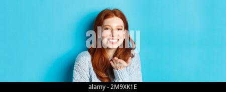 Lovely redhead female model smiling, sending air kiss at camera, standing against blue background Stock Photo