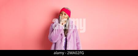 Fashion and shopping concept. Modern chinese senior woman shushing, telling a secret and looking cunning at camera, standing in trendy fake fur coat Stock Photo