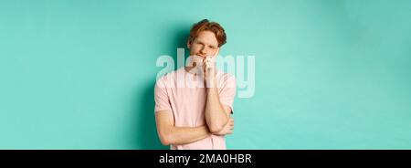 Bored handsome guy with red hair leaning on hand, looking tired and unamused at camera, standing in t-shirt against turquoise background Stock Photo