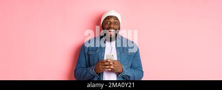 Technology and online shopping concept. Surprised young Black man using mobile phone, looking at camera amazed and happy after reading message, pink Stock Photo