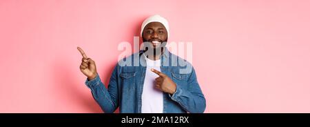 Happy african-american man smiling, pointing fingers right and showing promo, making announcement, standing over pink background Stock Photo