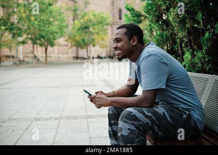 Man smiling while using mobile phone sitting on a bench outdoors. Stock Photo