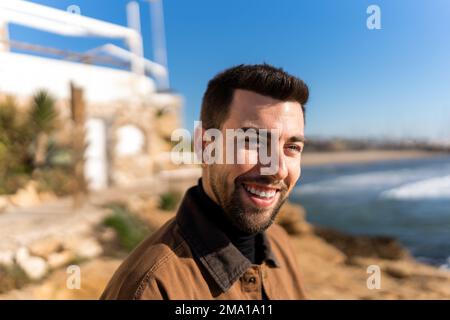 Positive Hispanic bearded male looking away on blurred background of aged building located on seashore in sunshine Stock Photo