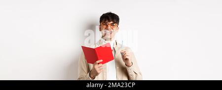 Cheerful young man in glasses laughing and taking notes, writing down in planner, holding pen and diary, standing on white background Stock Photo