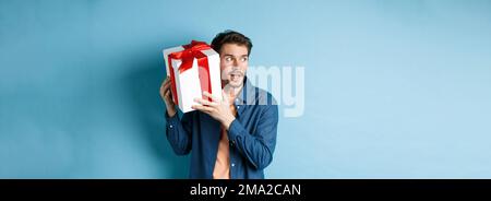 Valentines day. Curious boyfriend holding gift box near ear, trying to guess whats inside surprise present, standing over blue background Stock Photo