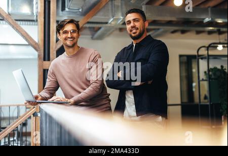 Male business professionals standing on an interior balcony and looking at the camera. Two business men working together in a tech startup. Stock Photo