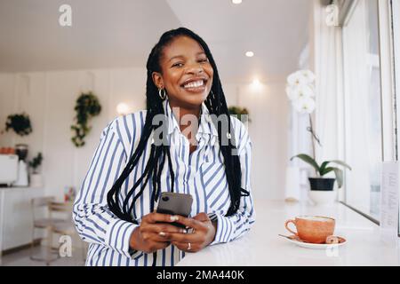Portrait of a young businesswoman smiling at the camera while holding a smartphone in a cafe. Happy young black businesswoman taking a coffee break in Stock Photo