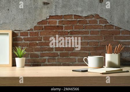 Stylish workplace with picture frame, cup of coffee, pencil holder and notebook on wooden table against brick wall Stock Photo