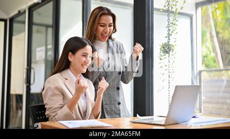 Two cheerful and excited millennial Asian businesswomen celebrating their project success together in the office, looking at laptop screen. Stock Photo