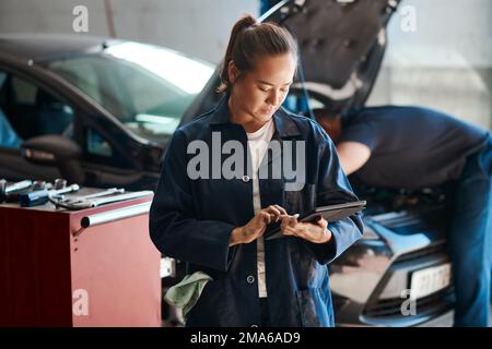 We use the latest diagnostic tools to fix problems. a female mechanic using a digital tablet while working in an auto repair shop. Stock Photo