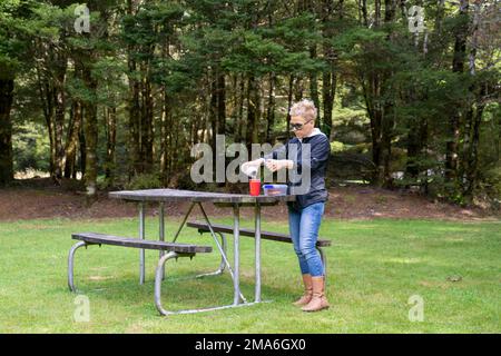 One blond white women in sunglasses is pouring coffee from thermos to the red cup on the picnic wooden table. Pleasant Flat Campsite. Haast Pass, West Stock Photo