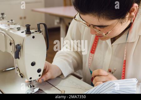 young seamstress woman in her workshop, sitting setting up the sewing machine, threading the needle to work on a design. Stock Photo