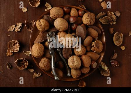 A variety of mixed nuts in a wooden bowl on a dark wooden tabletop with antique vintage nut crackers and shells scattered around Stock Photo
