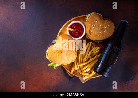 Valentine day Heart-shaped burgers. Two tasty cheeseburgers with french fries and beer bottles on dark table background. Idea for Valentine day dinner Stock Photo