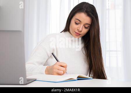 Focused young student working on a laptop, preparing for an exam and making notes. Stock Photo
