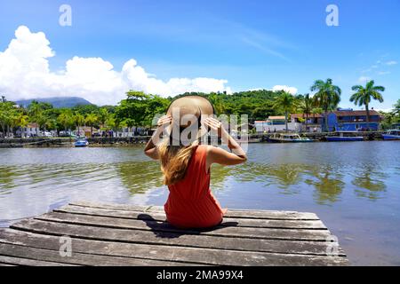 Tropical destination for Holidays. Beautiful young woman sitting on wooden pier enjoying relaxing sunny windy day in Brazil. Stock Photo