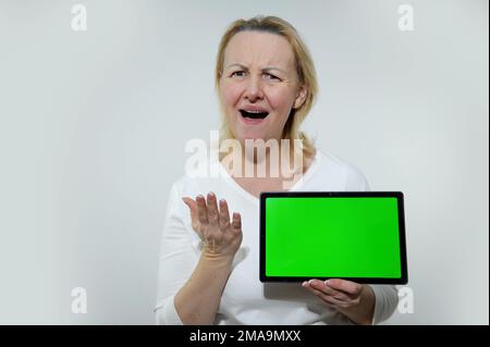 adult woman with tablet in her hands is very surprised she does not like what she sees on it shows a green chromakey screen on a white screen Stock Photo
