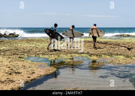 Three young surfers walk on the rocks towards Pedra do Sal beach in Salvador, Brazil. Stock Photo