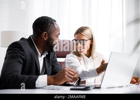 Two people analyze documents on a laptop while sitting at a table in the office. Business partners at work in the office are discussing projects Stock Photo