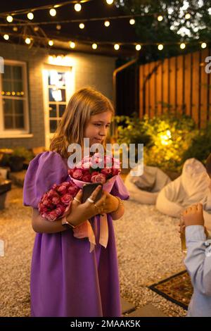 Cute teen girl with rose flowers. Child playing in summer garden. Kids gardening. Children play outdoors. Blond female with flower bouquet for birthda Stock Photo