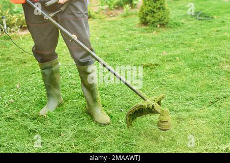 Man or worker mowing the green grass. Gardener mows weeds with electric or petrol lawn trimmer in city park or backyard. Gardening care tools and equi Stock Photo
