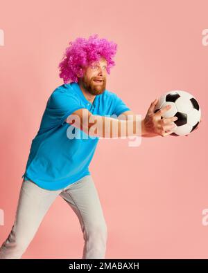 Emotional football fan, young caucasian man in funny wig watching soccer match and expressing emotions over pink background. Sport, fans, football Stock Photo