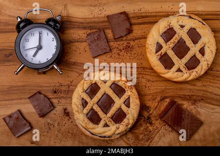 Two chocolate tarts sprinkled with cocoa powder, chocolate squares and an alarm clock that strikes seven taken from above Stock Photo