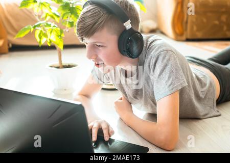boy in wireless gamer headphones lies on floor and plays online games. child learns programs remotely at home in winter garden near flower in pot Stock Photo