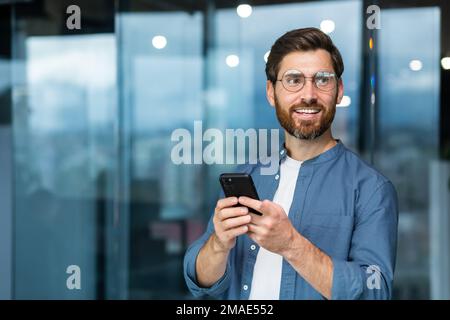 Portrait of a young handsome male student in glasses standing in the campus, office. He holds the phone in his hands, smiles, looks to the side. Stock Photo