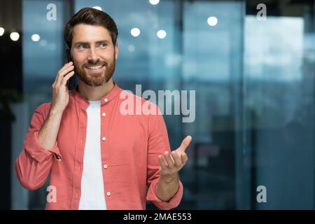 Portrait of a young handsome man standing and talking on the phone indoors, office. Stock Photo