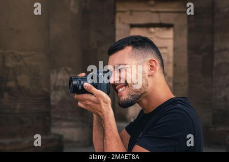 Caucasian young man with dark hair looking into camera viewfinder and photographing while smiling in a temple. Stock Photo