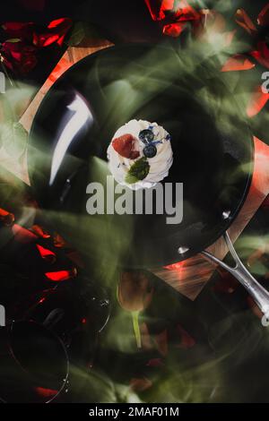 black frying pan with a pavlova cake and scattered red rose petals on a wooden board with a black background with a glass of red wine and lit candles Stock Photo