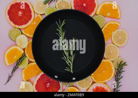 black frying pan with a sprig of rosemary in it on top of sliced citrus herbs. Oranges, grapefruit, lemon, lime on a pink background. flat lay Stock Photo