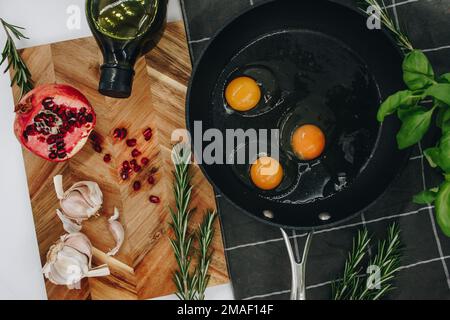 black frying pan on a black towel with three raw eggs in it next to a wooden cutting board with a red pomegranate on it. Stock Photo