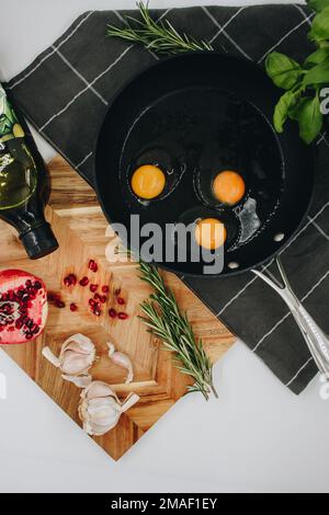 black frying pan on a black towel with three raw eggs in it next to a wooden cutting board with a red pomegranate on it. Stock Photo