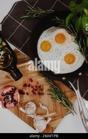 black frying pan on a black towel with three fried egg yolks in it next to a wooden cutting board with a red pomegranate on it. Stock Photo
