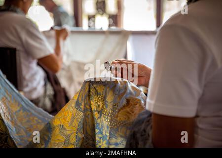 Indonesian male and female artists drawing batik on blue fabric in a touristic atelier. Close up shot from the back. Stock Photo