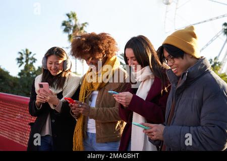 Four friends of diverse nationalities gathered outdoors, standing in a row, using mobile phones. Stock Photo